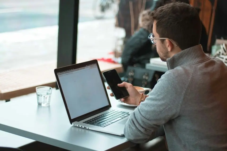 A man at a laptop checking his phone by a window