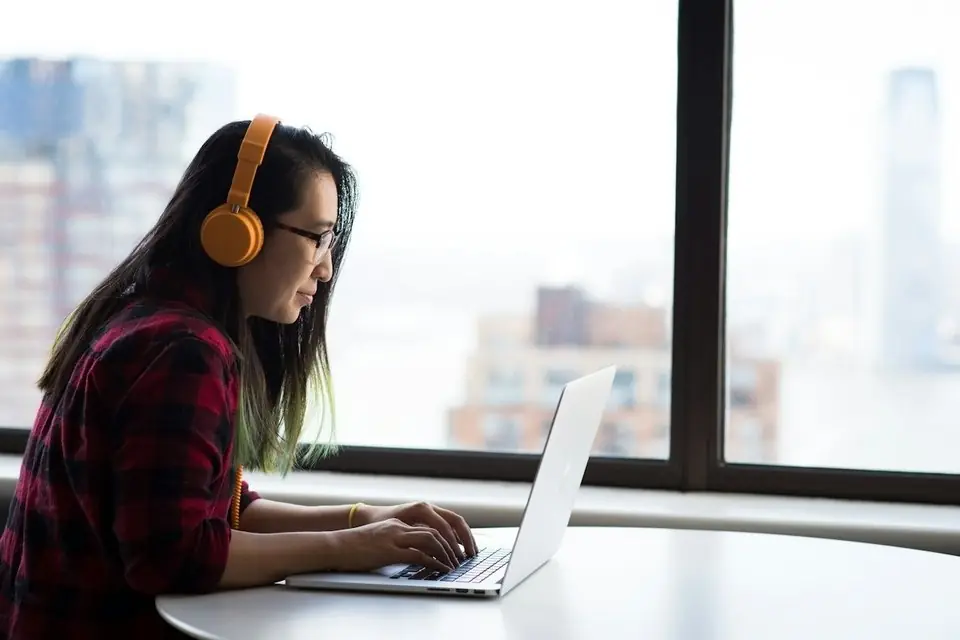 A woman in headphones working at a laptop by a window